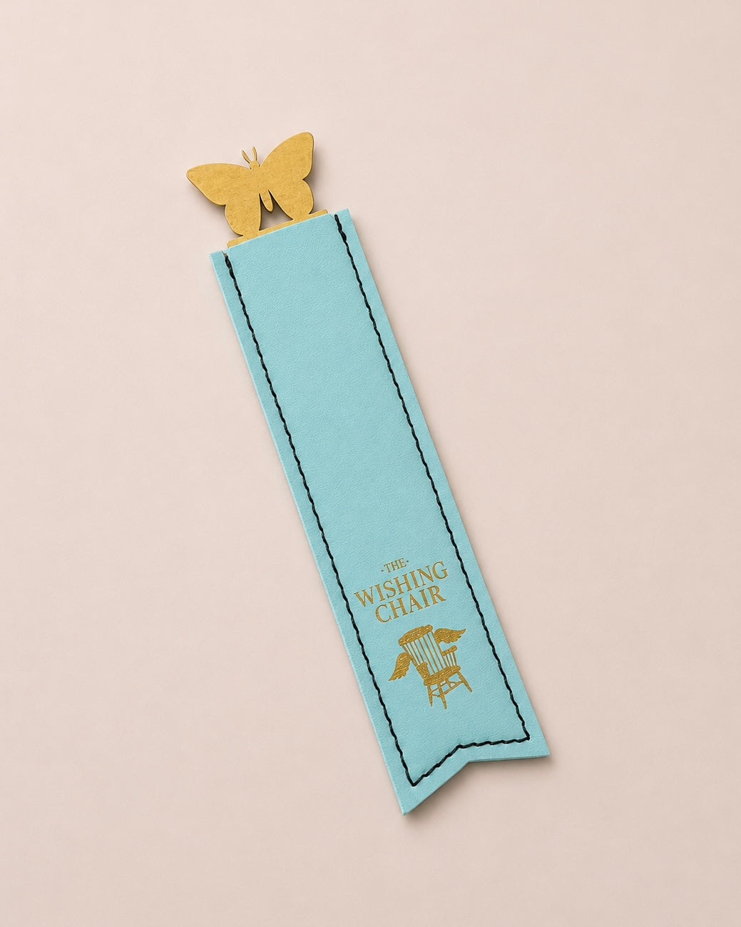 A light blue bookmark shaped like a ribbon bookmark with a gold butterfly clip at the top and gold foil text reading 'THE WISHING CHAIR' with a chair graphic, shown on a pale pink background.