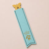 A light blue bookmark shaped like a ribbon bookmark with a gold butterfly clip at the top and gold foil text reading 'THE WISHING CHAIR' with a chair graphic, shown on a pale pink background.